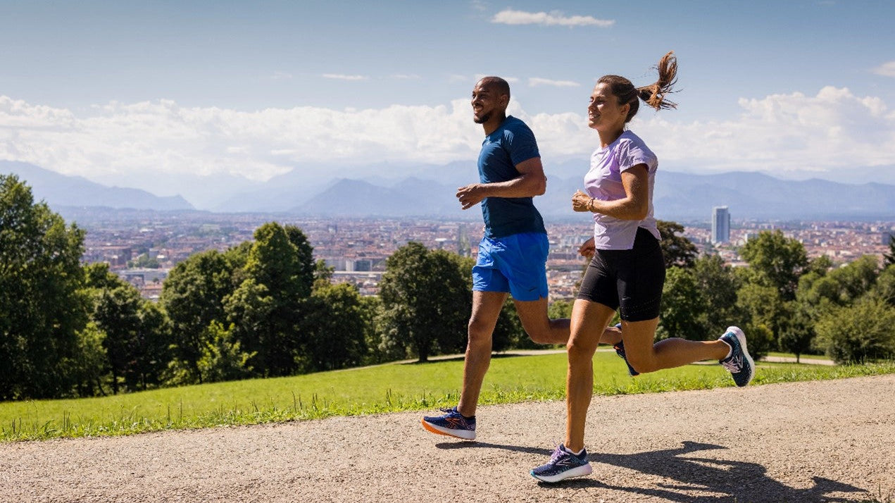 Ein Mann und eine Frau laufen in BROOKS-Ausrüstung bei gutem Wetter auf einem Weg.