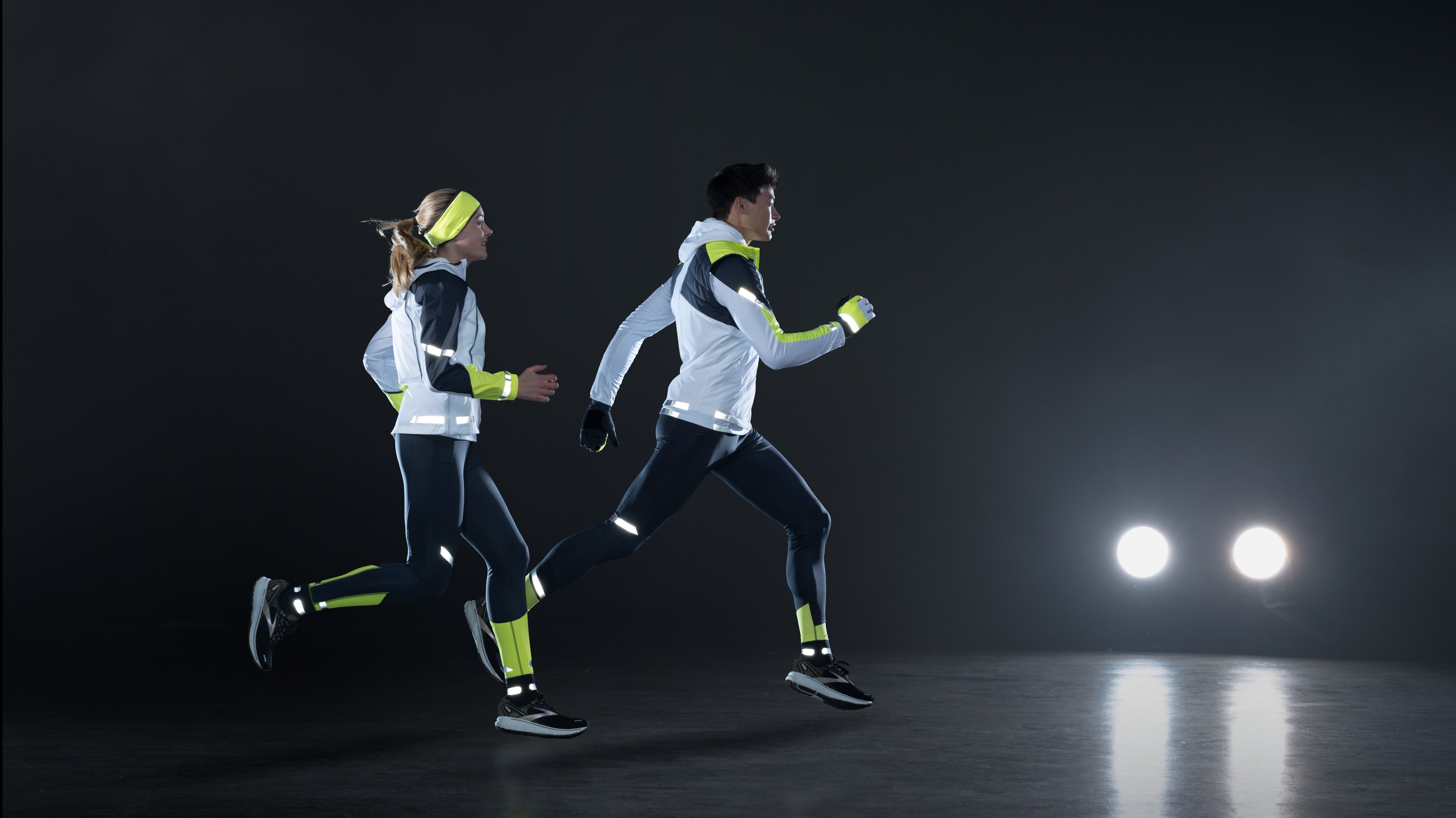 2 Personen laufen mit BROOKS Run Visible Bekleidung durch die Dunkelheit. In der Ferne sind 2 Scheinwerfer eines Autos zu sehen.