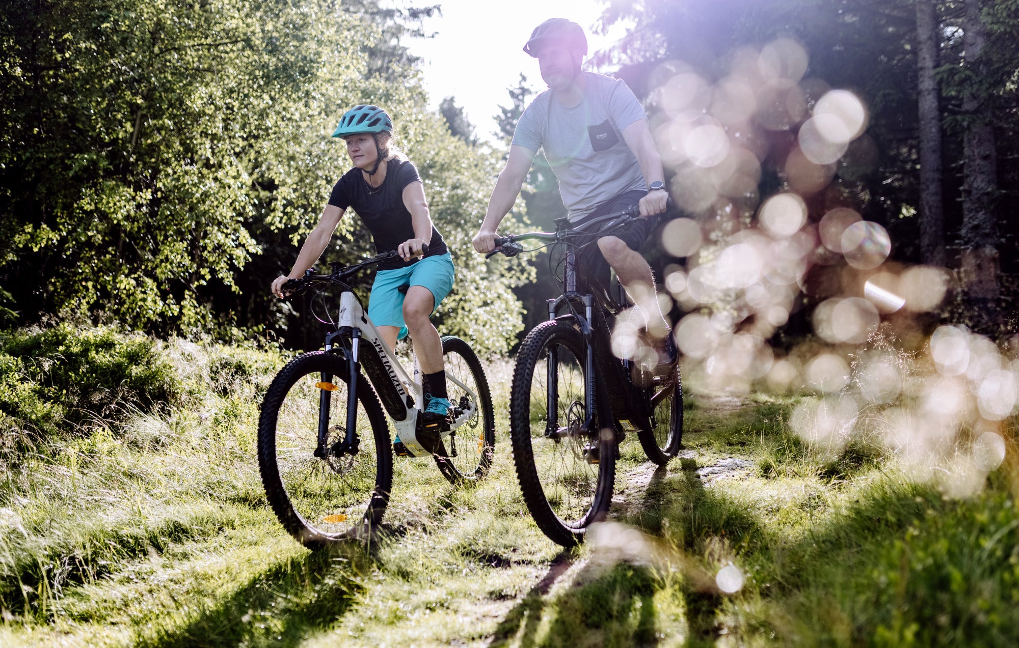 Eine Frau und ein Mann fahren mit sommerlicher Bekleidung mit SPECIALIZED Fahrrädern durch den Wald.