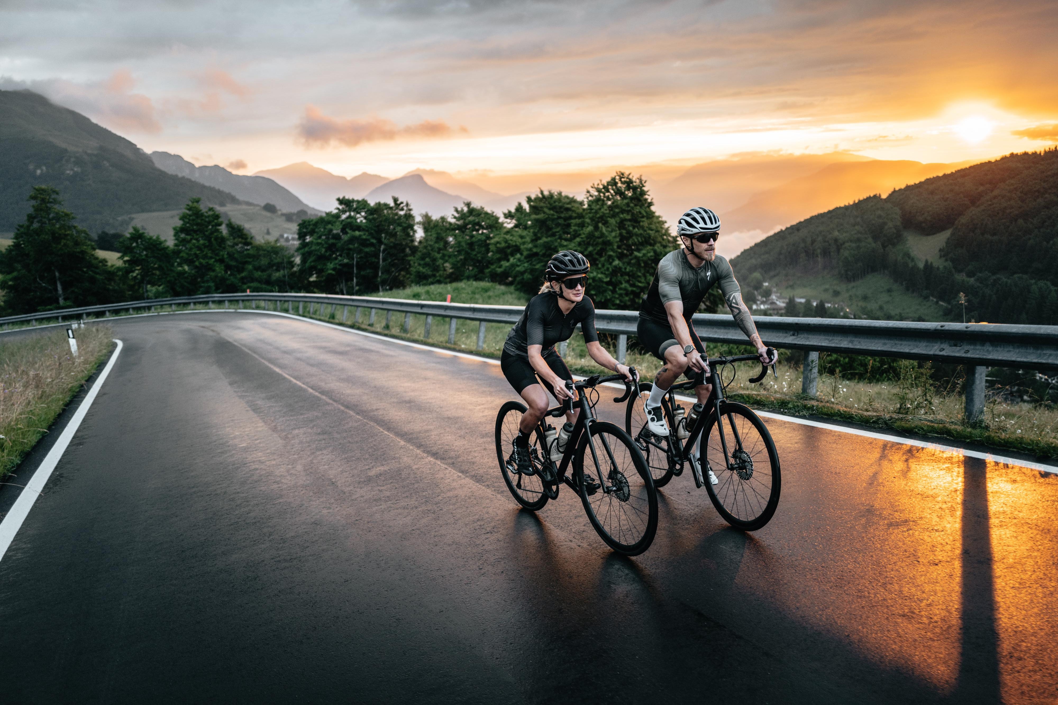 1 Frau und ein Mann fahren mit Rennrädern auf einer Landstraße den Berg im Sonnenuntergang hoch.