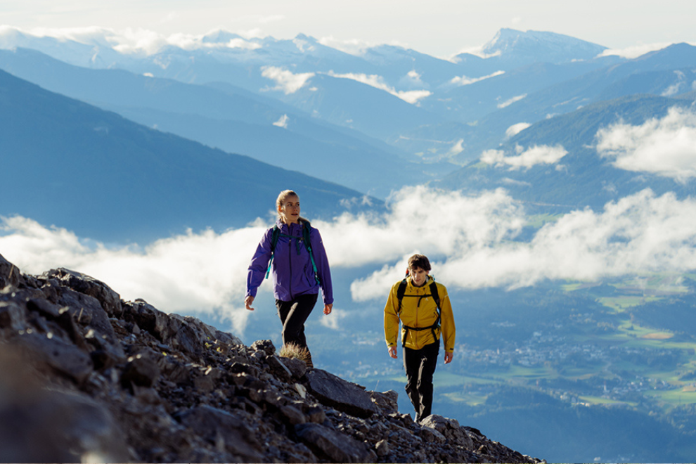 Eine Frau und ein Mann besteigen einen Berg. Im Hintergrund sieht man weitere Berge und das Tal.