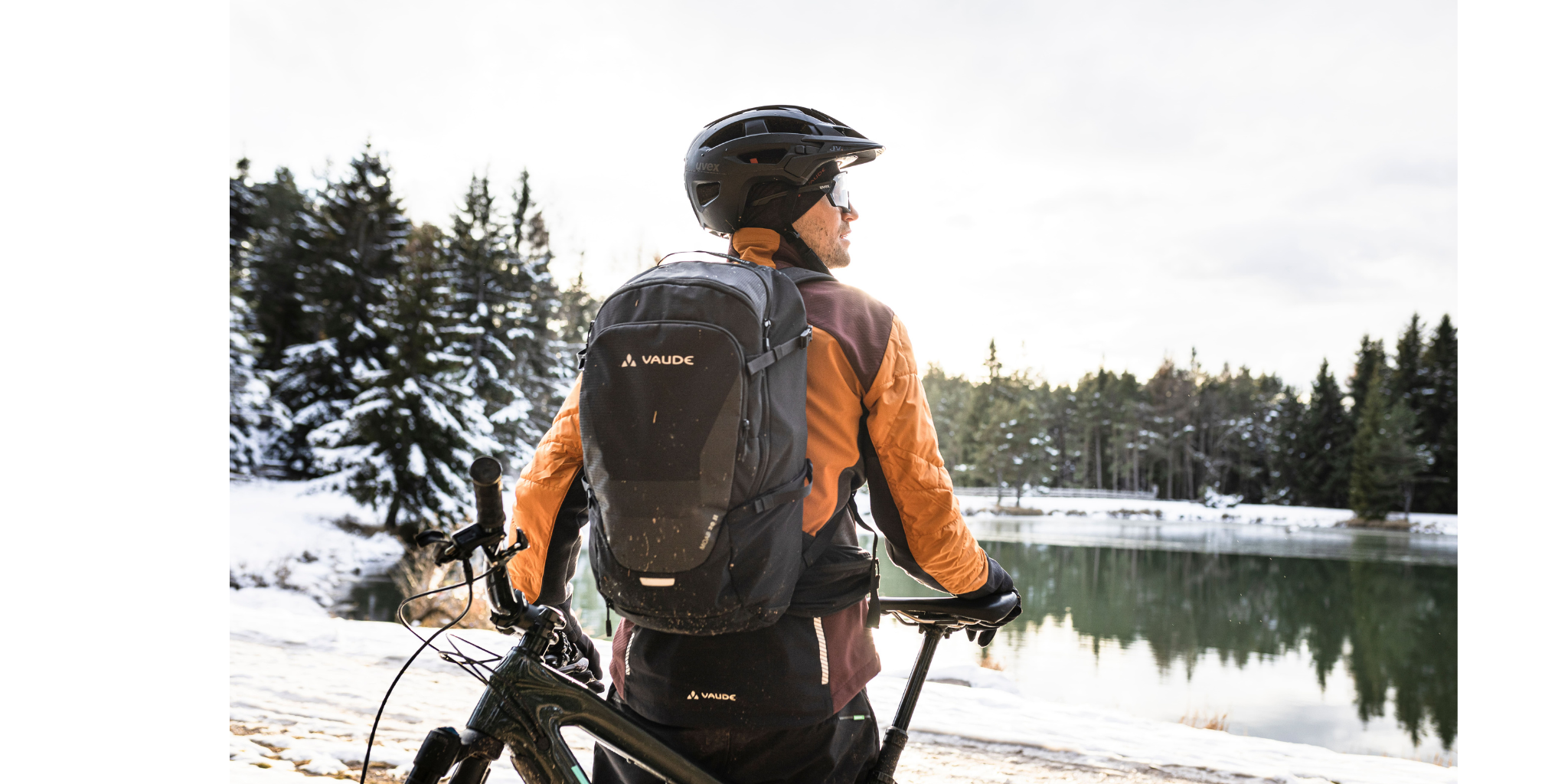 Ein Mann hat sein Fahrrad in der Hand und blickt in die Ferne. Dabei ist er im Winter im Wald an einem See.