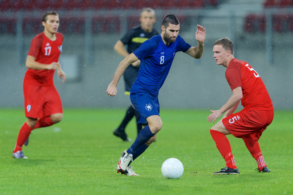 Ein Fußballer in einem blauen Trikot dribbelt mit dem Ball durch 2 Gegenspieler in roten Trikots.