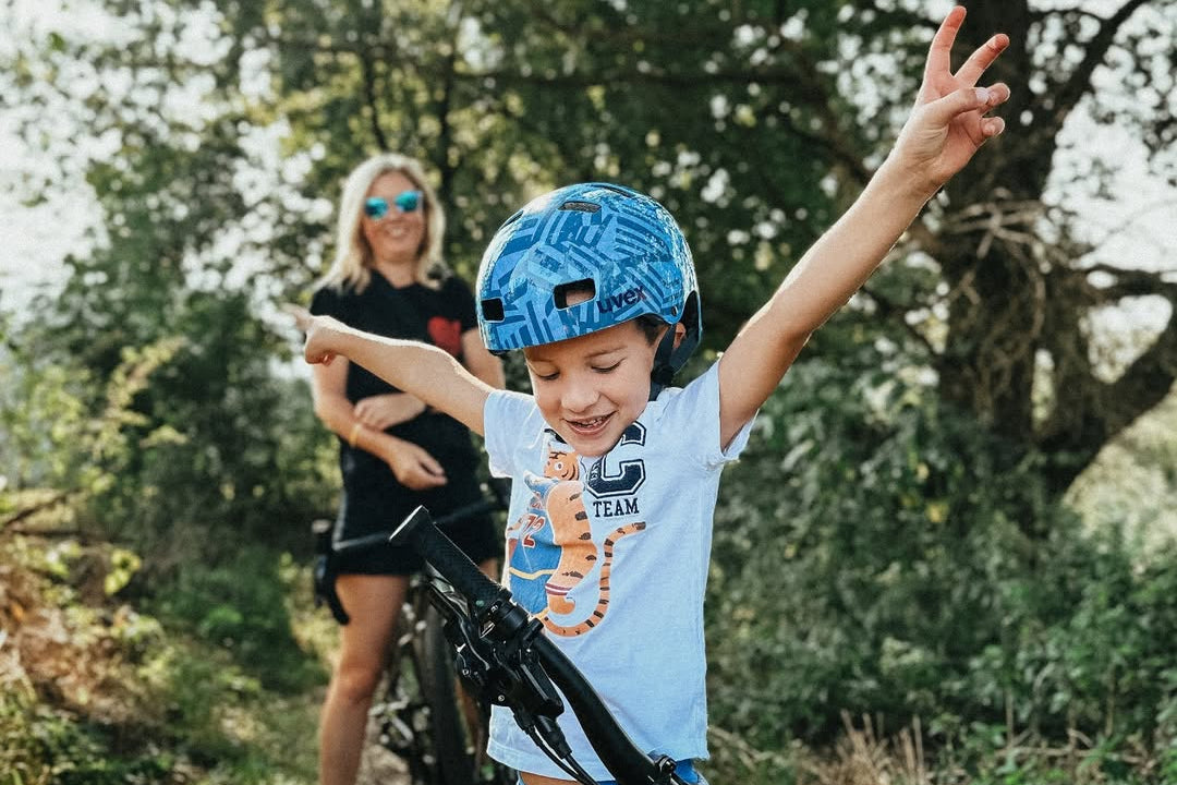 Kind mit Woom Fahrrad im Vordergrund mit einem Helm auf. Im Hintergrund eine blonde Mutter in der grünen Natur mit Wiese und einem Baum