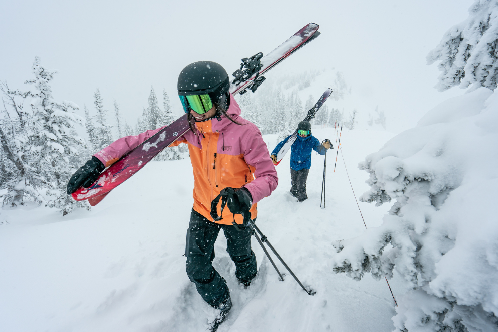2 Personen laufen in Ski-Bekleidung mit Skiern durch einen leichten Schneeregen.
