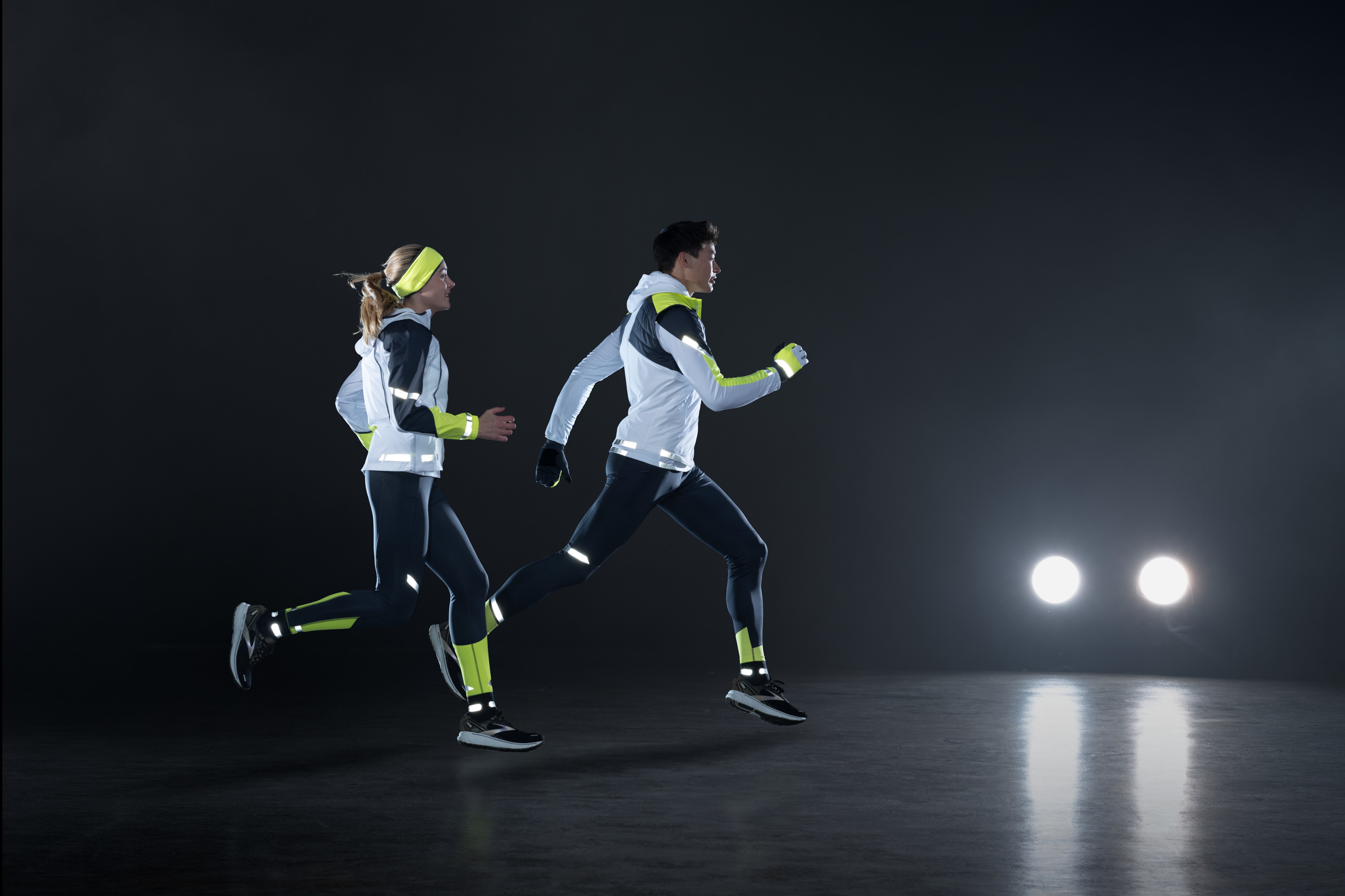 2 Personen laufen mit BROOKS Run Visible Bekleidung durch die Dunkelheit. In der Ferne sind 2 Scheinwerfer eines Autos zu sehen.