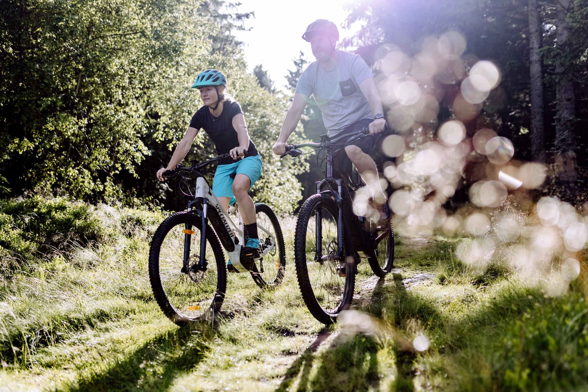 Eine Frau und ein Mann fahren mit sommerlicher Bekleidung mit SPECIALIZED Fahrrädern durch den Wald.