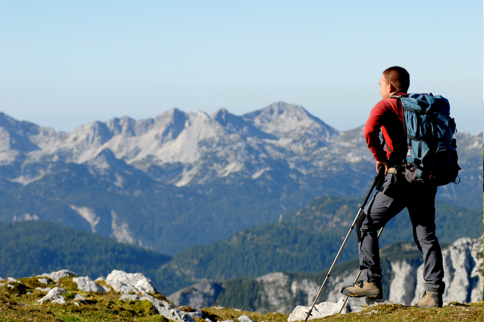 1 Mann steht auf einem Berg und schaut in die Ferne. Im Hintergrund ist das Tal und die Berglandschaft zu sehen.