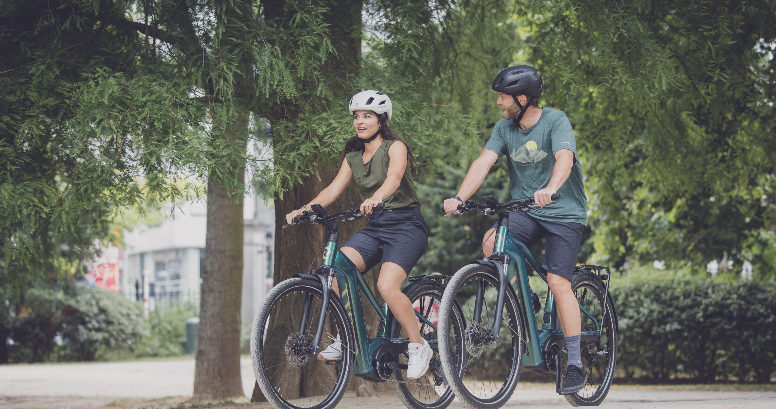 Eine Frau und ein Mann fahren auf SCOTT-E-Bikes durch einen Park.