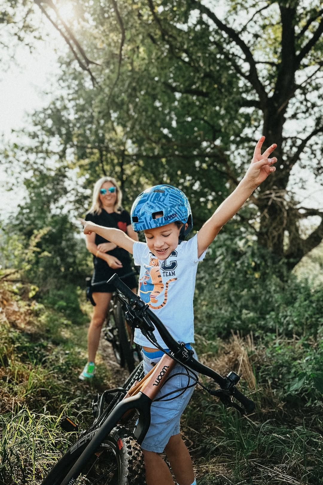 Kind mit Woom Fahrrad im Vordergrund mit einem Helm auf. Im Hintergrund eine blonde Mutter in der grünen Natur mit Wiese und einem Baum 