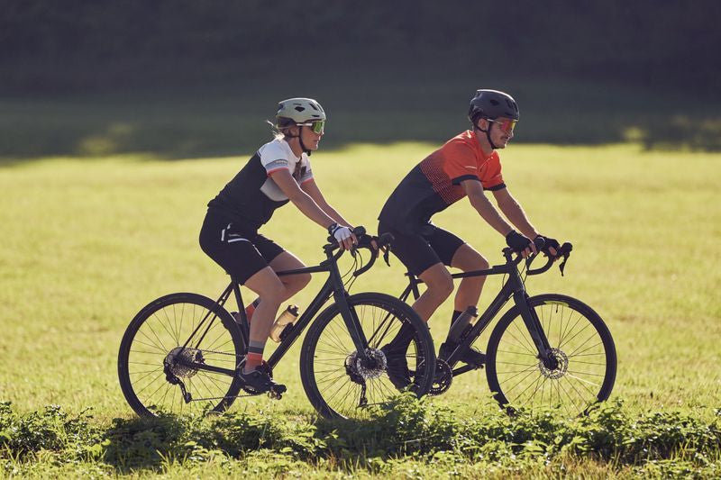 Eine Frau und ein Mann fahren in Fahrradtrikots mit Gravelbikes. Im Hintergrund ist eine grüne Wiese zu sehen.